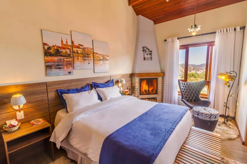 Um quarto aconchegante com uma cama de casal, lareira, poltrona, obras de arte na parede e uma grande janela com vista para o exterior, na Pousada Suíça Mineira, para representar pousadas em Monte Verde.