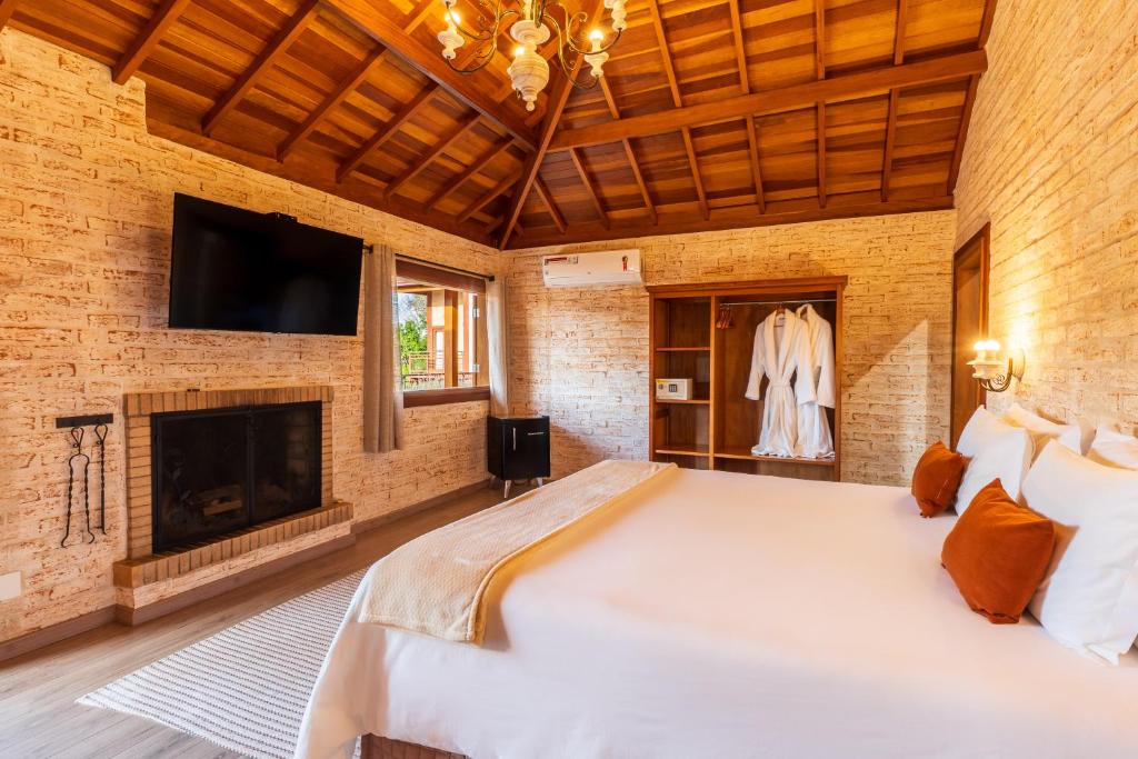 Quarto com uma cama grande, lareira, TV na parede, armário aberto e janela; teto de madeira e paredes de tijolos, na Pousada Vilage de Minas, para representar pousadas em Monte Verde.