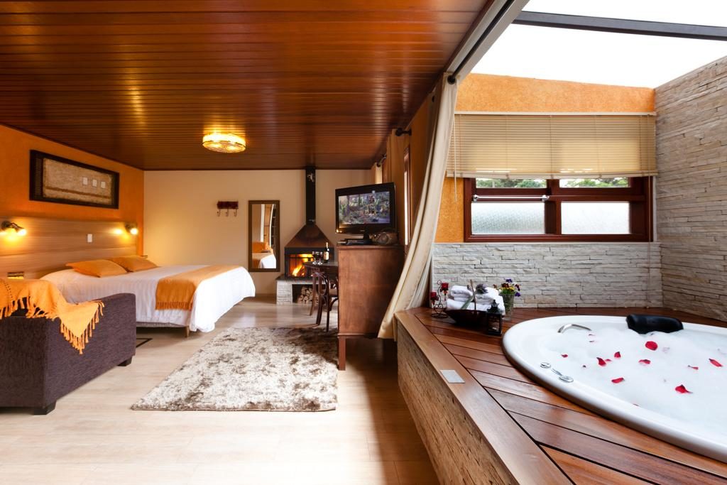 Quarto da Pousadas Águia Dourada com uma cama de casal à esquerda, uma lareira ao fundo do quarto e uma banheira de hidromassagem arredonda à direita da foto, para representar pousadas em Monte Verde.