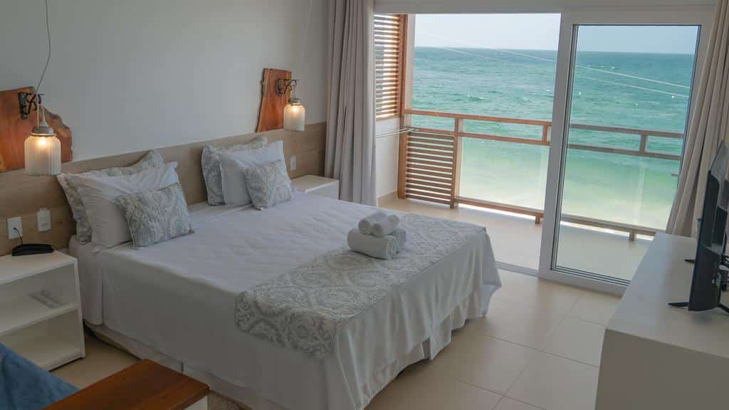 Um quarto moderno na Pousada Dona Moça com uma cama grande, roupa de cama branca, varanda com vista para o mar e luz natural das portas de vidro deslizantes, para representar pousadas em Morro de São Paulo.