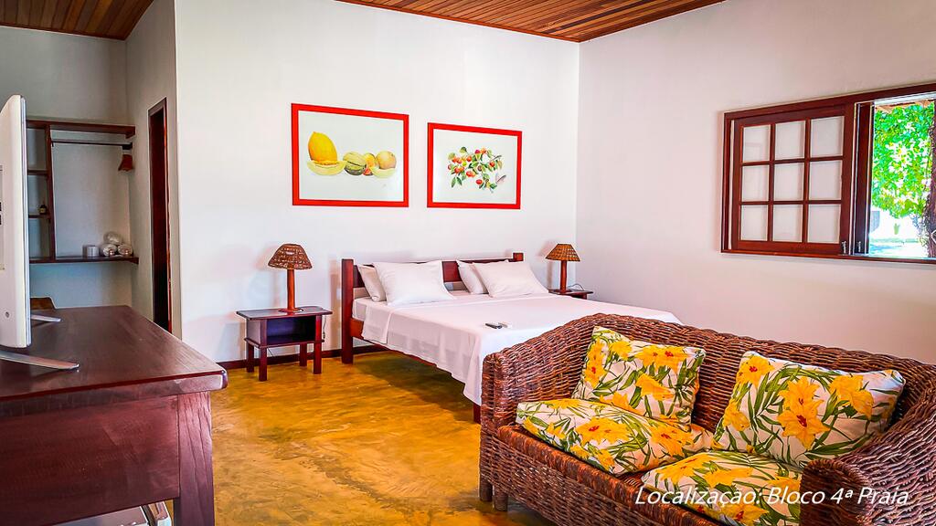 Quarto bem iluminado na Pousada Caeira, com uma cama de casal, sofá de vime, mesas laterais, arte de frutas emoldurada e uma janela com vista para o exterior, para representar pousadas em Morro de São Paulo.