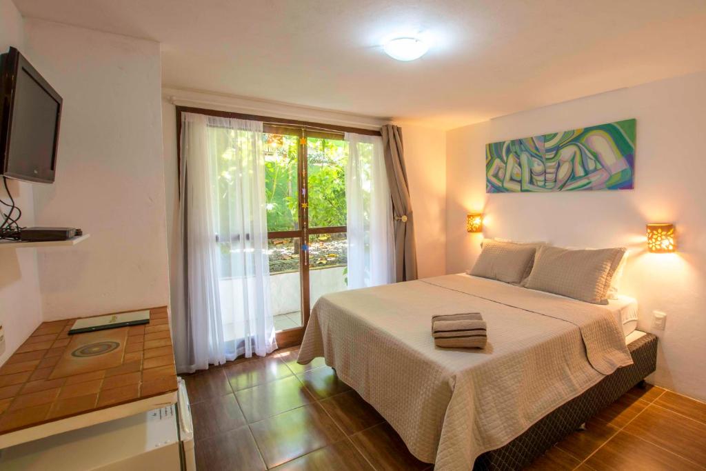 Um quarto simples na Pousada Borboleta com uma cama de casal, toalhas dobradas, arte na parede, TV, fogão e portas de vidro deslizantes para um pátio com vegetação, para representar pousadas em Morro de São Paulo.