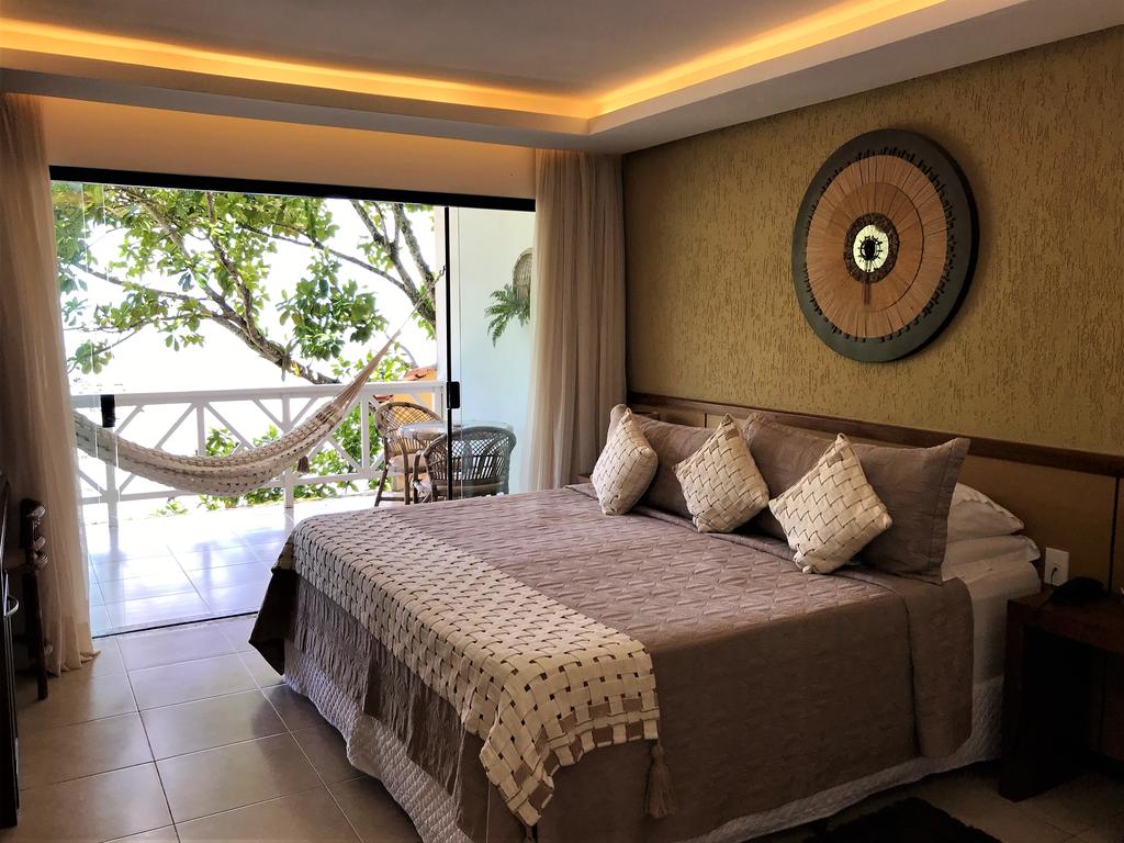 Um quarto com uma cama bem arrumada, travesseiros decorativos e uma rede em uma varanda com vista para as árvores, na Passárgada Pousada e Restaurante, para representar pousadas em Morro de São Paulo.
