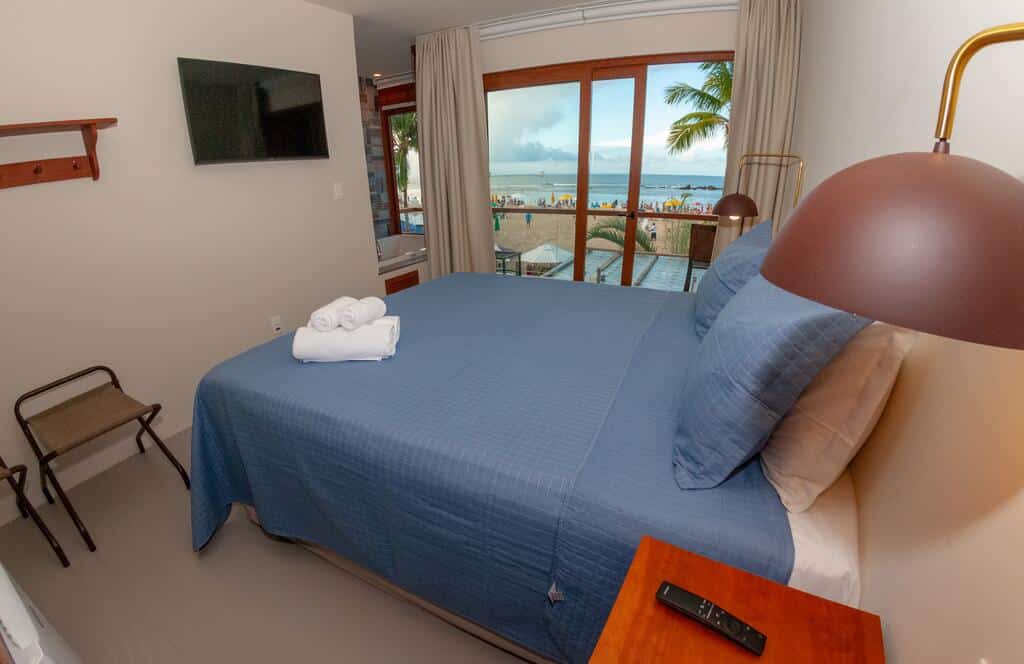 Quarto no Club do Balanço Pousada e Restaurante com colcha azul, TV na parede, janela com vista para a praia e toalhas dobradas sobre a cama, para representar pousadas em Morro de São Paulo.