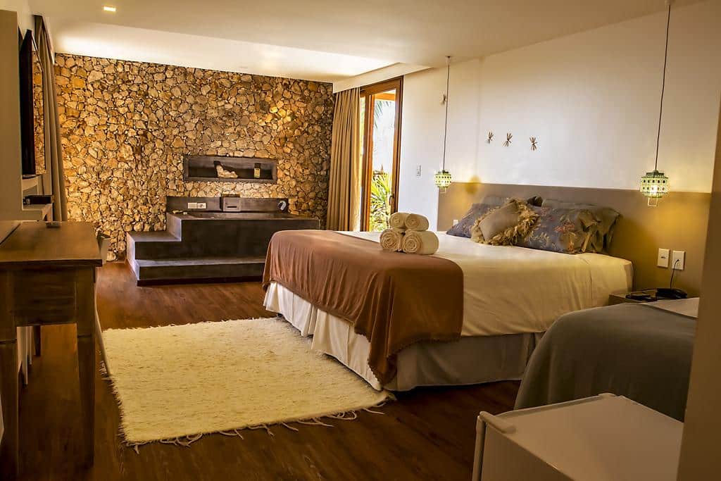 Quarto com parede de pedra, cama de casal, roupa de cama marrom e branca, tapete branco e luzes pendentes, na Pousada Minha Louca Paixão, para representar pousadas em Morro de São Paulo.