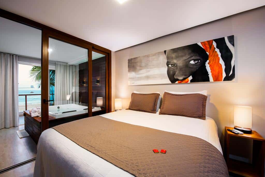 Quarto moderno na Pousada Bahia Tambor com uma cama de casal, luminárias de cabeceira, jacuzzi, varanda e um retrato artístico acima da cama, para representar pousadas em Morro de São Paulo.