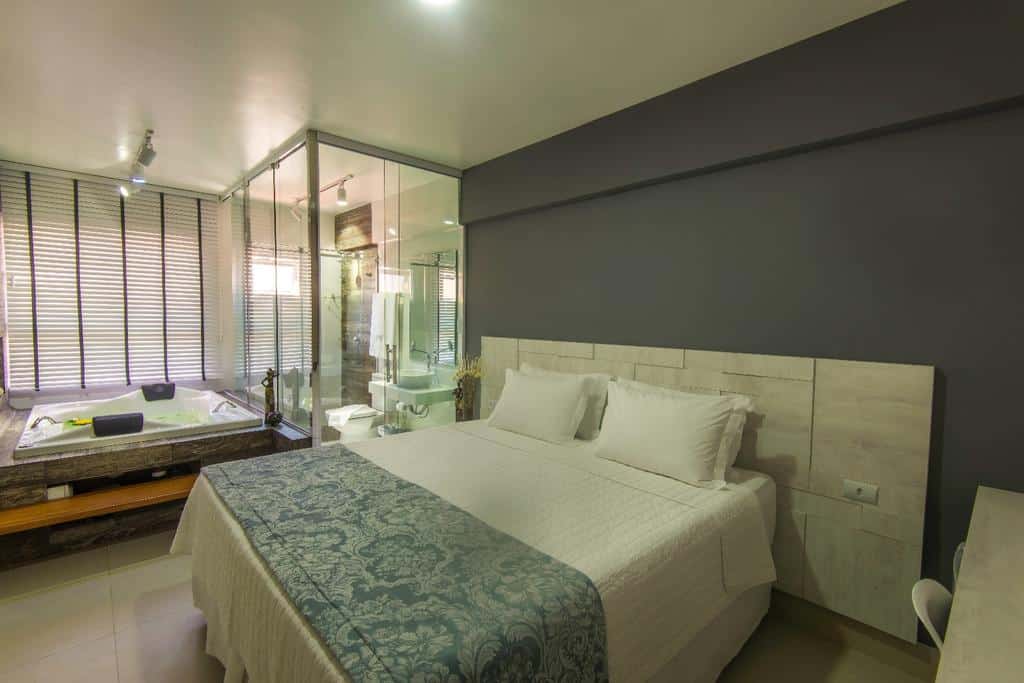 Quarto moderno na Cristal Pousada com uma cama de casal, banheiro com paredes de vidro e uma jacuzzi ao lado de grandes janelas com persianas, para representar pousadas em Morro de São Paulo.