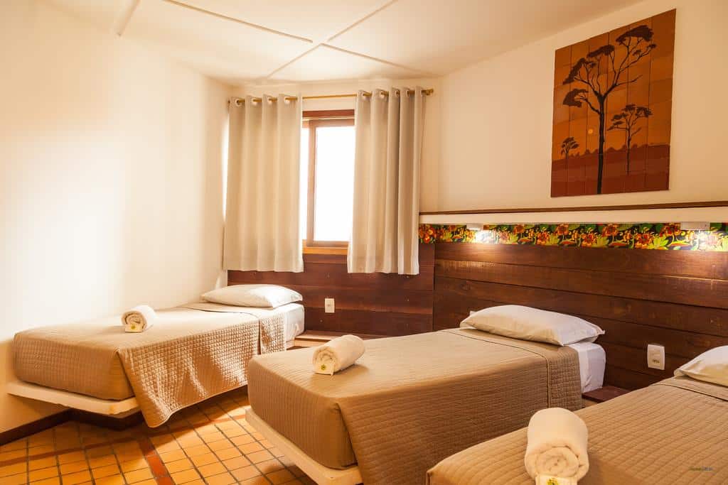 Três camas de solteiro com roupa de cama bege em um quarto bem iluminado, uma janela com cortinas e uma arte de árvore na parede, na Pousada Bahia Brasil, para representar pousadas em Morro de São Paulo.