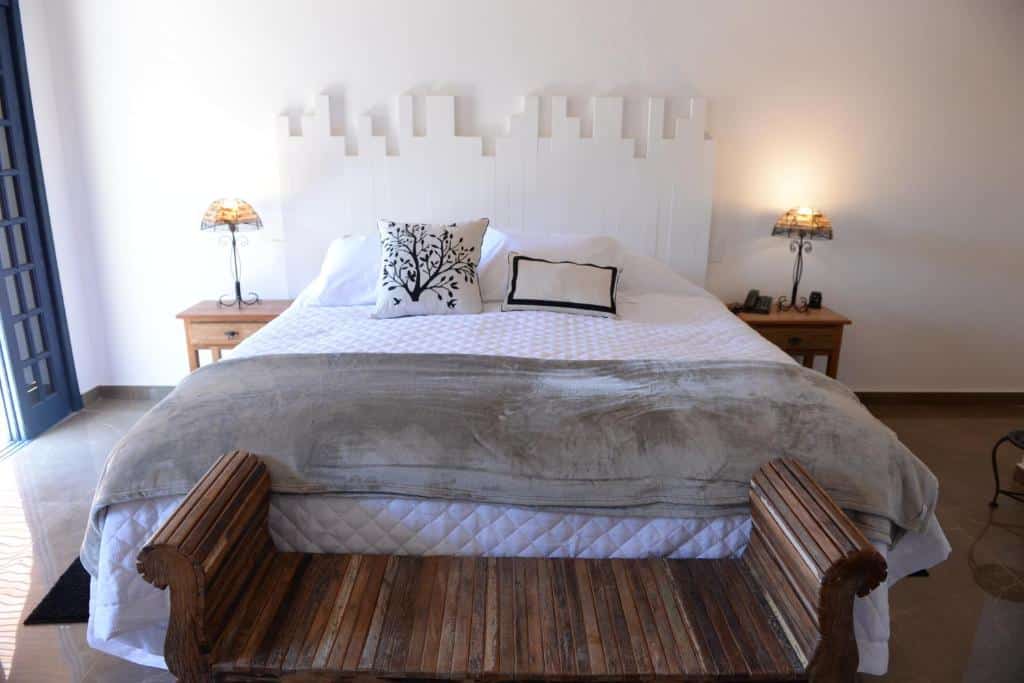 Quarto da Le Garden Pousada Boutique, uma das pousadas em Penedo, com uma cama bem arrumada com roupa de cama branca, duas mesas de cabeceira com abajures e um banco de madeira aos pés da cama.