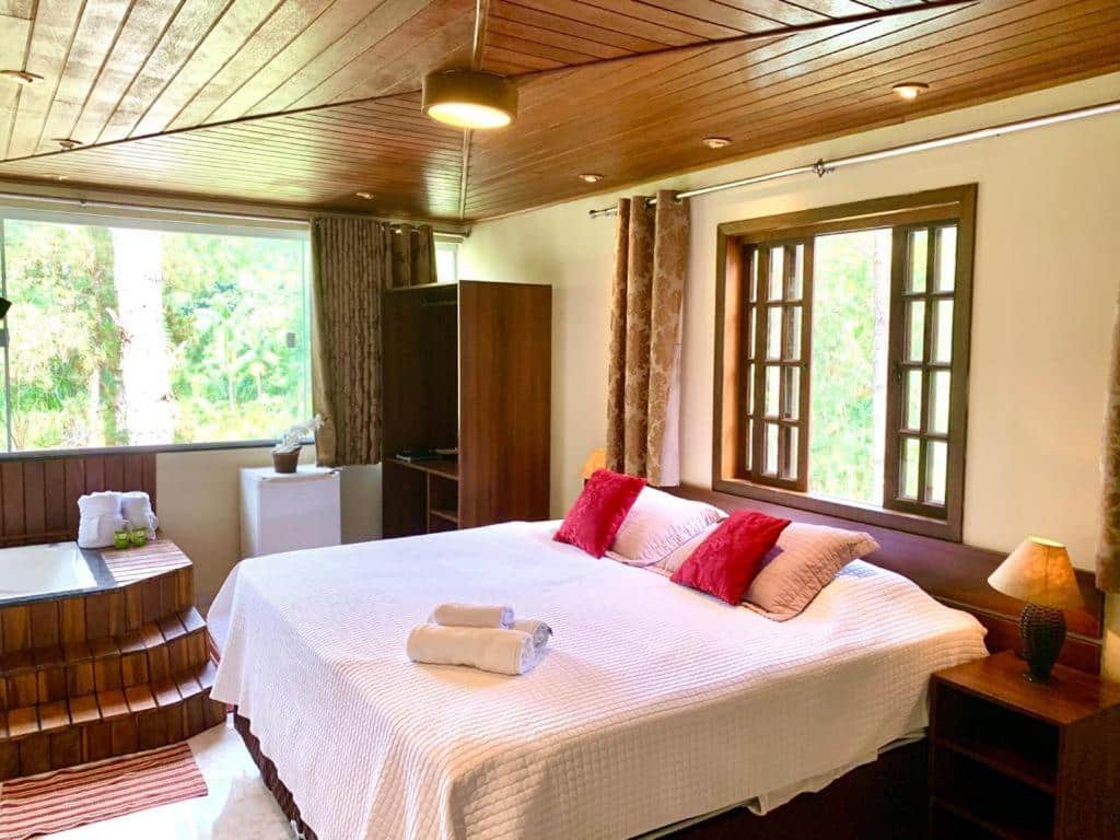 Quarto da Recanto de Moriá, uma das pousadas em Penedo, com uma cama grande, roupa de cama branca, travesseiros vermelhos, uma janela, uma banheira de hidromassagem e um pequeno frigobar próximo ao canto.