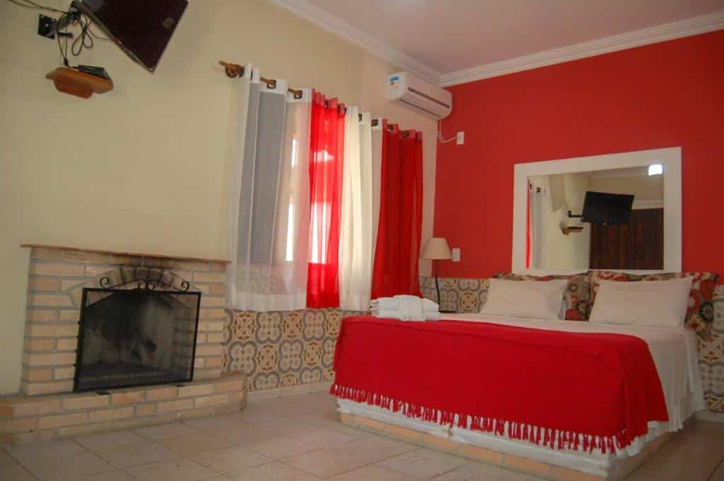 Quarto da Pousada do Sol, uma das pousadas em Penedo, com uma cama coberta com lençóis vermelhos e brancos, uma lareira, um espelho de parede, uma TV e cortinas vermelhas e brancas.
