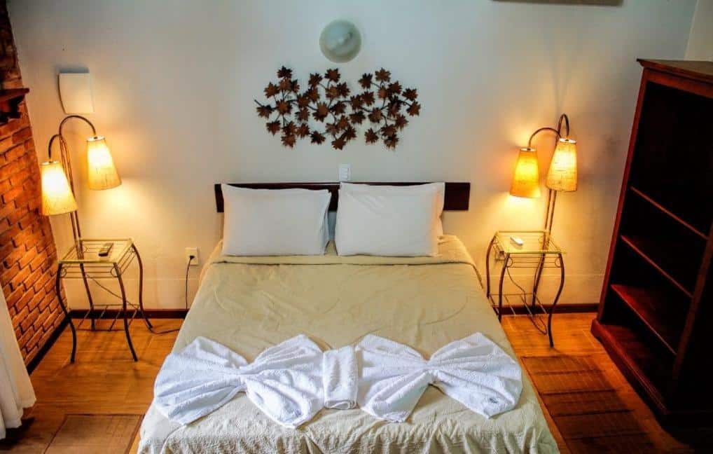 Quarto da Pousada Viking Penedo, com uma cama bem arrumada com dois travesseiros brancos, toalhas dobradas sobre o cobertor e abajures nas mesas laterais em um quarto aconchegante.