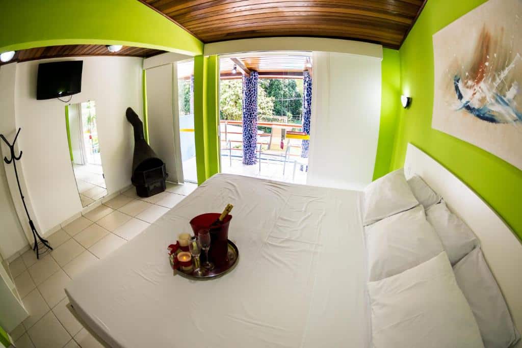Quarto moderno da Pousada Suarez, uma das opções de pousadas em Penedo, com cama de casal, parede com detalhes verdes, TV, espelho e uma bandeja com bebidas sobre a cama.