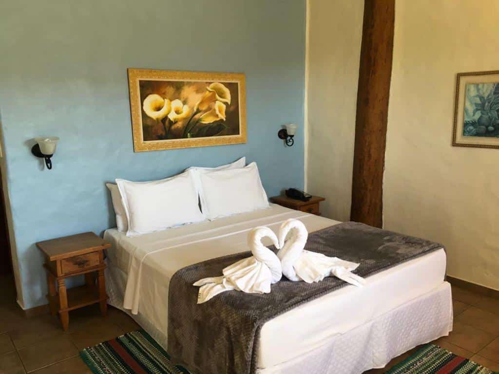 Quarto da Pousada Santa Fé de Penedo com uma cama bem arrumada com lençóis brancos, cisnes de toalha, duas mesinhas de cabeceira e obras de arte florais na parede.