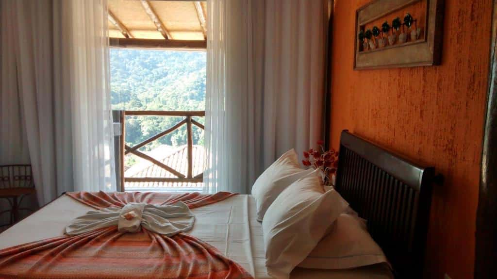 Quarto da Pousada Terraço Penedo com uma cama bem arrumada, com toalhas dobradas, fica de frente para uma janela com vista para a montanha, cortinas brancas e uma parede laranja de destaque.