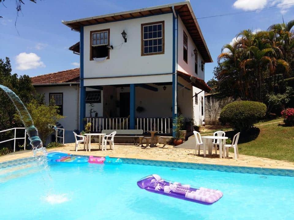 Piscina da Pousada Casarão De Minas, com uma pequena queda d'água, algumas boias coloridas, a pousada logo atrás e um pequeno jardim