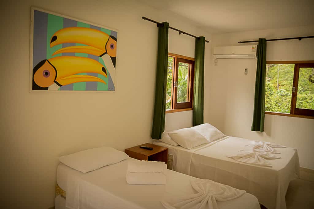 Um quarto simples com duas camas de solteiro, cortinas verdes, duas janelas e uma pintura de tucano na parede branca, na Pousada Sol da Trindade.
