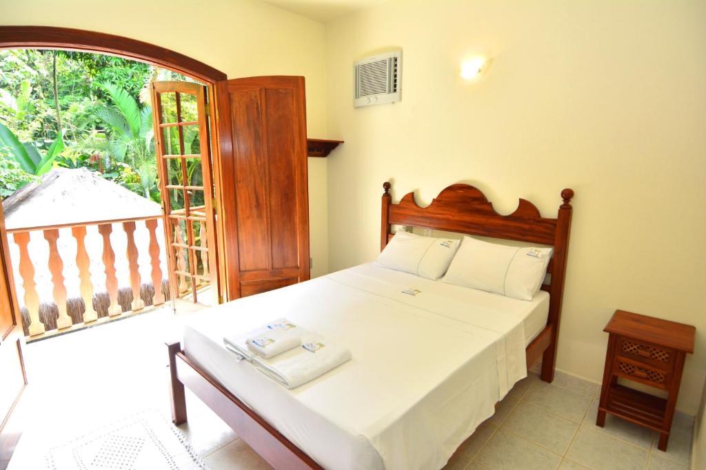 Um pequeno quarto com cama de madeira, roupa de cama branca, mesa de cabeceira e porta aberta que dá para uma varanda com vegetação externa, na Pousada Dois Irmãos, para representar pousadas em Trindade.