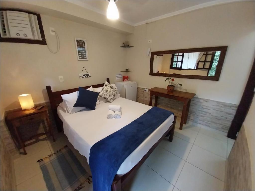 Cama bem arrumada com roupa de cama branca e detalhes em azul em um quarto pequeno com escrivaninha, frigobar e espelho de parede, na Pousada Refúgio do Pirata, para representar pousadas em Trindade.