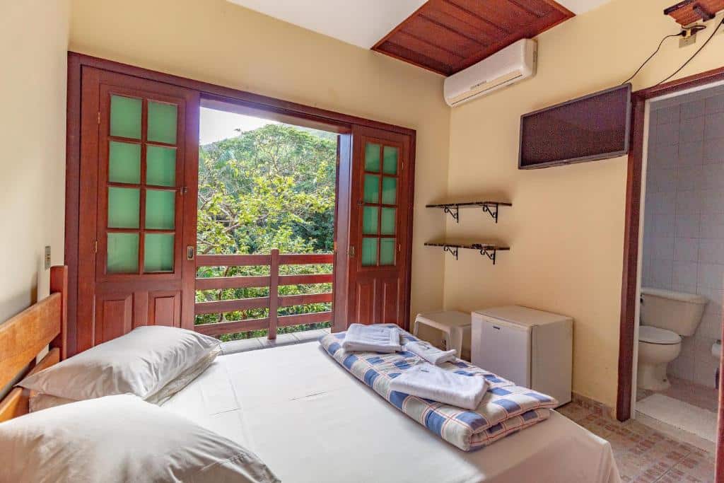 Quarto pequeno com cama de casal, toalhas dobradas, portas abertas para a varanda, ar-condicionado, TV e vista para a vegetação externa, na Pousada do Marquinhos, para representar pousadas em Trindade.