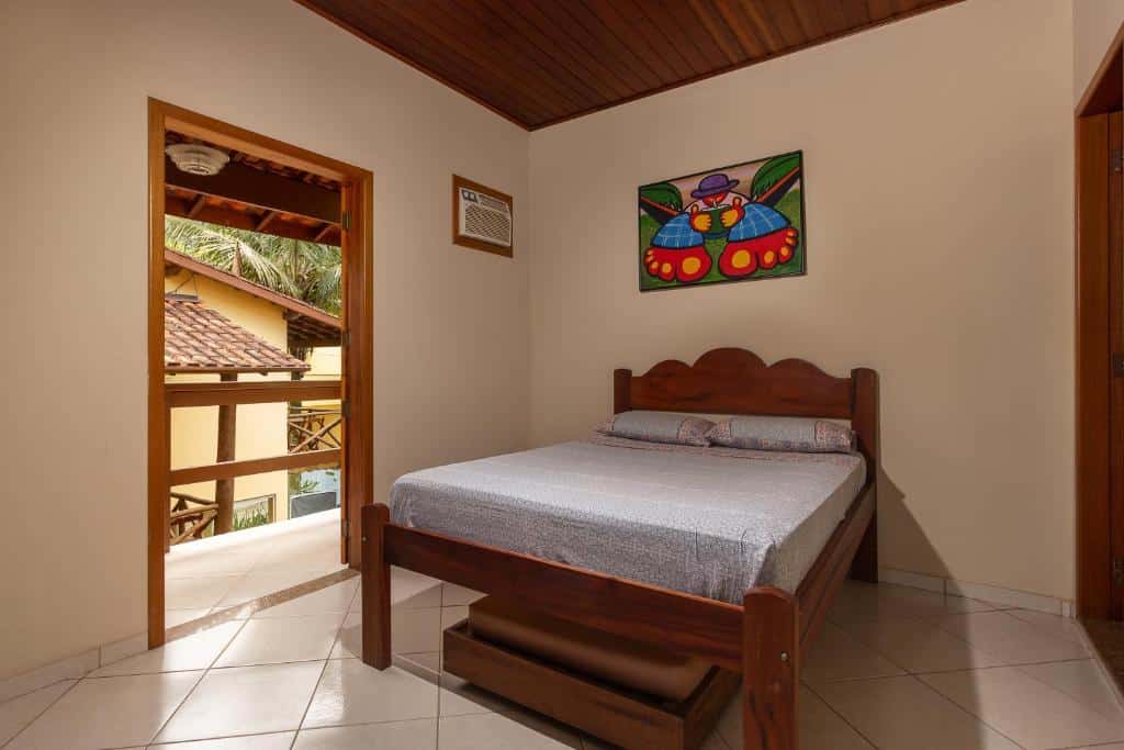 Um pequeno quarto com cama de madeira, roupa de cama cinza, piso de ladrilho, obras de arte acima da cama e uma porta aberta para uma varanda, na Pousada Cabeça do Indio, para representar pousadas em Trindade.