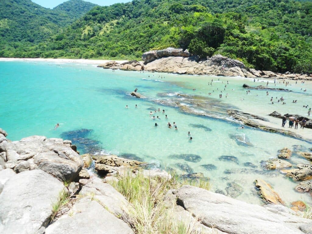 Pessoas nadando em água azul-turquesa clara perto da costa rochosa e de colinas verdes sob um céu claro em uma praia tropical, na Praia do Meio, em Trindade.