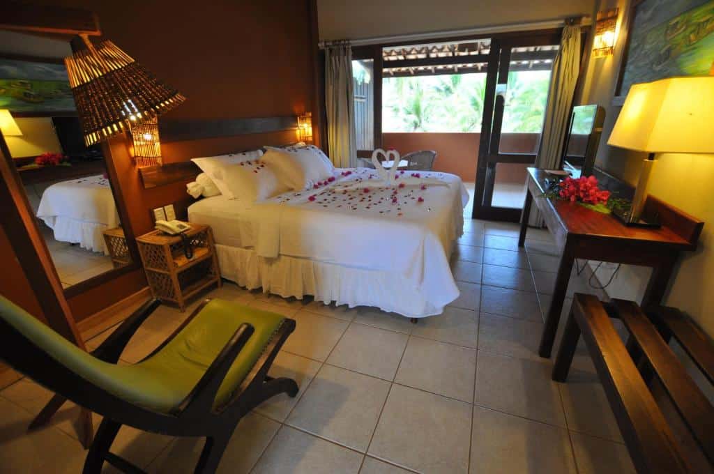 Quarto na Pousada Jambo em Itacimirim com cama de casal, pétalas de rosa, escrivaninha, cadeira, luminária, espelho grande e varanda com vista para a vegetação, para rperesentar pousadas na Linha Verde na Bahia.