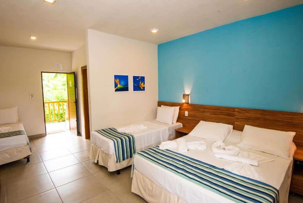 Quarto na Pousada Tropicarim em Itacimirim, com três camas, colchões listrados, parede com detalhes em azul, arte na parede e uma porta aberta para uma varanda, para rperesentar pousadas na Linha Verde na Bahia.