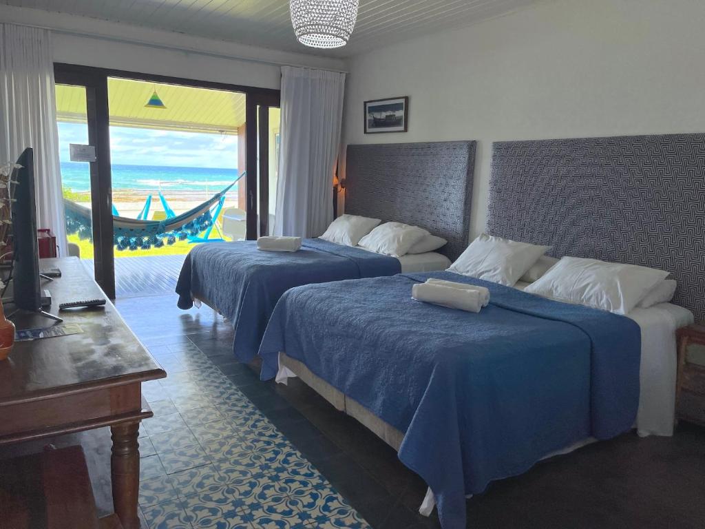 Um quarto na Pousada a Capela em Arembepe, com duas camas, roupa de cama azul, uma escrivaninha e uma rede em um pátio de frente para o oceano, para representar pousadas na Linha Verde na Bahia.
