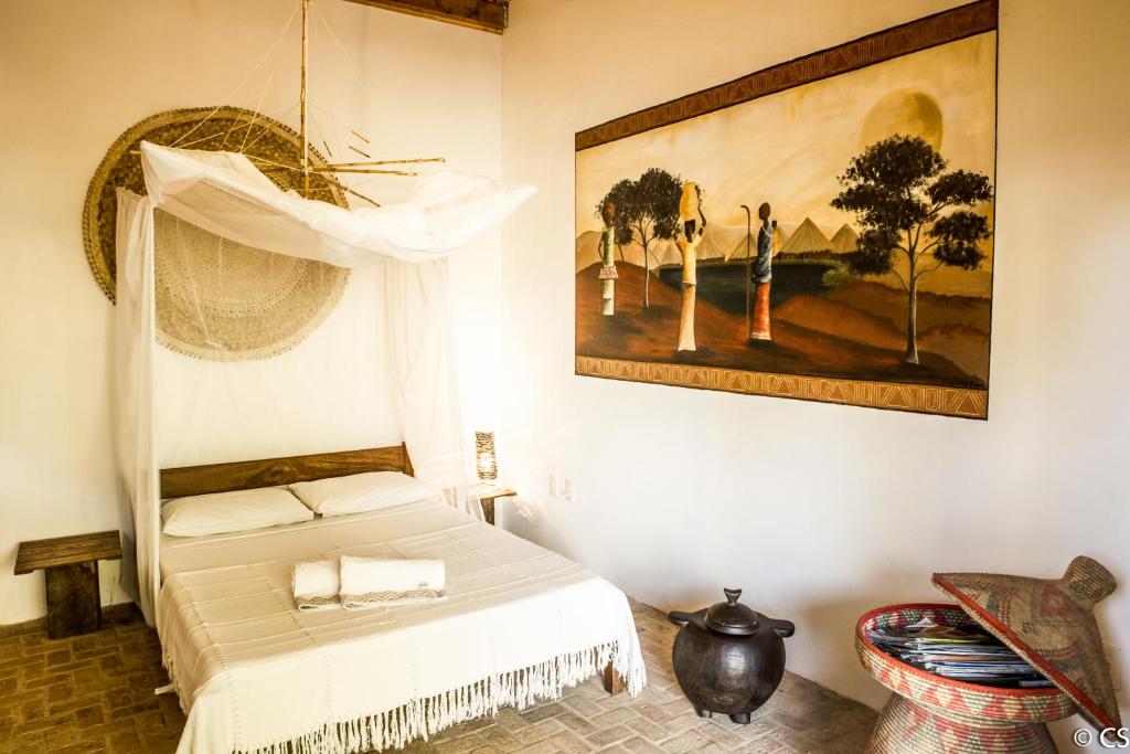 Uma cama bem arrumada com um dossel branco, decoração em tecido, obras de arte africanas na parede e cestas no chão em um quarto bem iluminado, na Pousada Too Cool em Diogo, para representar pousadas na Linha Verde na Bahia.