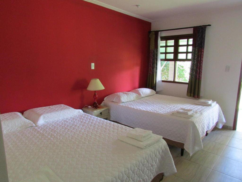 Quarto na Pousada Ipitanga IV em Lauro de Freitas, com parede vermelha, duas camas de casal com roupa de cama branca, mesa de cabeceira, abajur, janela e toalhas dobradas, para representar pousadas na Linha Verde na Bahia.