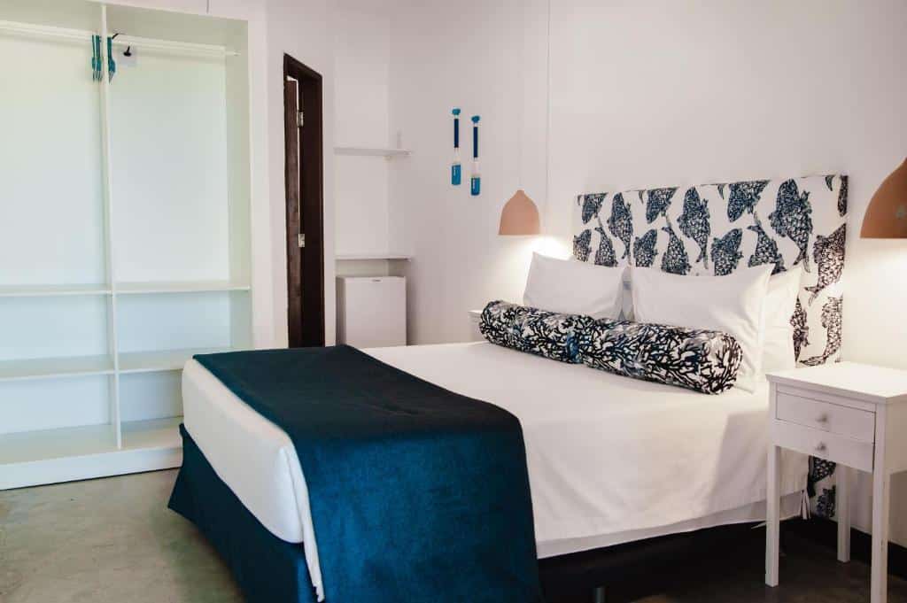 Uma cama bem arrumada com cabeceira estampada, roupa de cama branca, cobertor azul e um criado-mudo em um quarto minimalista, na Pousada Aldeola by Slaviero Hotéis em Baixio, para representar pousadas na Linha Verde na Bahia.