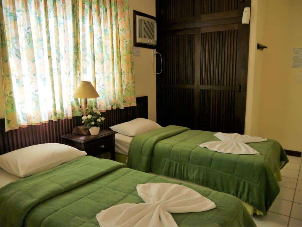Um quarto com duas camas de solteiro cobertas com cobertores verdes, cortinas florais e uma mesa de cabeceira com uma lâmpada e flores, na Grimms Pousada de Contos na Costa do Sauípe, para representar pousadas na Linha Verde na Bahia.