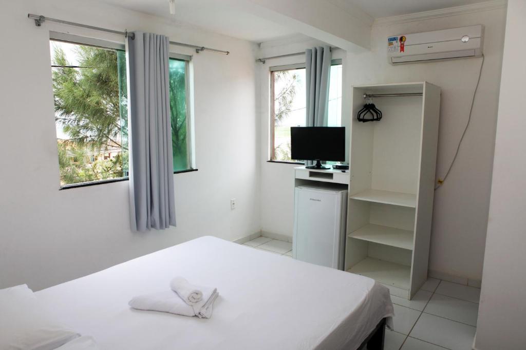 Um quarto simples com uma cama, duas janelas, uma TV, frigobar, armário e ar-condicionado. As toalhas ficam sobre a cama, no Hotel Pousada Oasis em Conde, para representar pousadas na Linha Verde na Bahia.