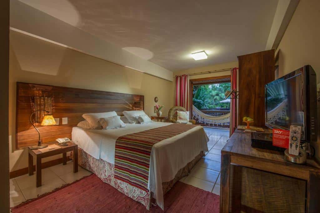Quarto no Refúgio da Vila na Praia do Forte, com uma cama king-size, móveis de madeira, TV e uma varanda com rede com vista para a vegetação, para representar pousadas na Linha Verde na Bahia.
