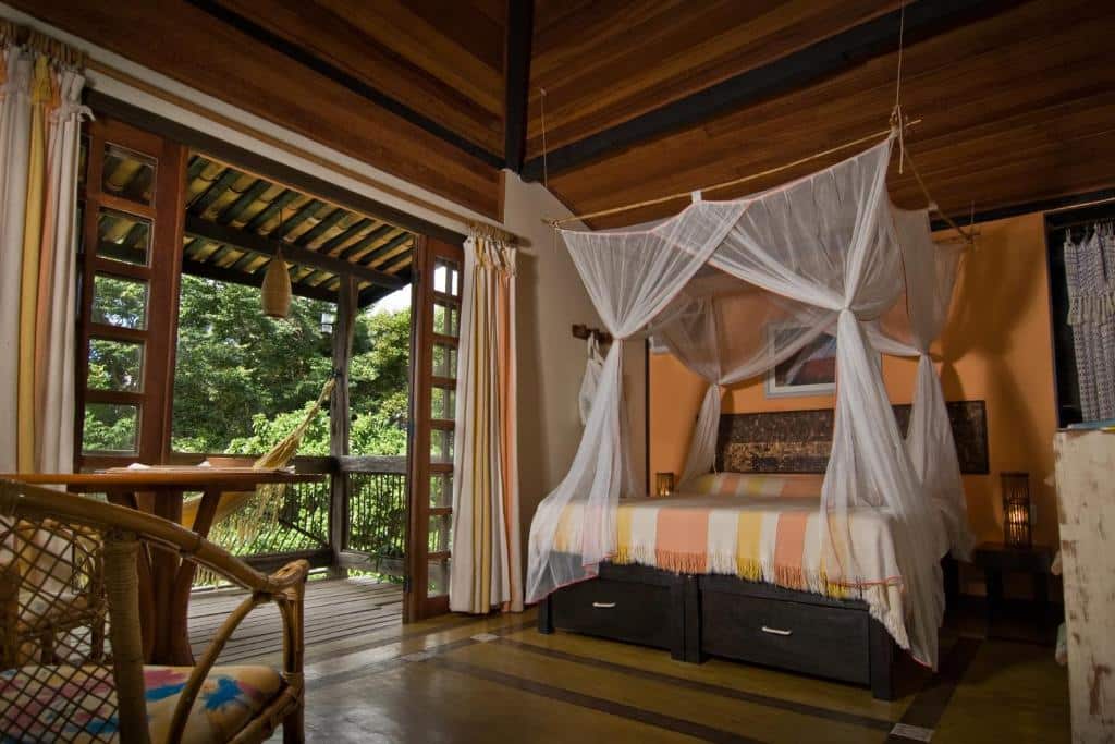 Uma cama de dossel com mosquiteiro fica em um quarto com teto de madeira, ao lado de portas abertas que levam a uma varanda, na Vilangelim Eco-Pousada em Imbassaí, para representar pousadas na Linha Verde na Bahia.
