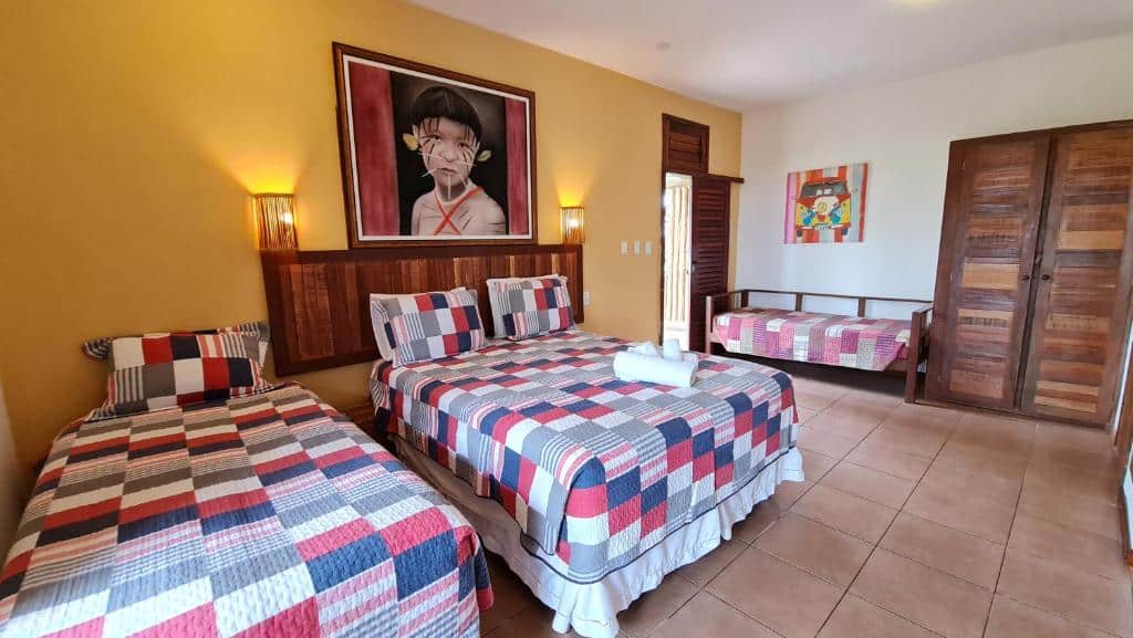 Quarto com uma cama de casal e duas camas de solteiro, todas com roupa de cama de retalhos e arte colorida nas paredes amarelas e brancas, na Pousada Praia das Ondas – Pé na areia em Itacimirim, para representar pousadas na Linha Verde na Bahia.