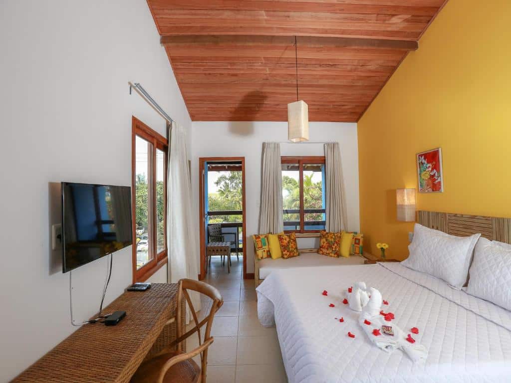 Quarto na Pousada Paraíso do Forte na Praia do Forte, com uma cama grande, TV, escrivaninha, área de estar e uma varanda com vegetação tropical visível do lado de fora, para representar pousadas na Linha Verde na Bahia.
