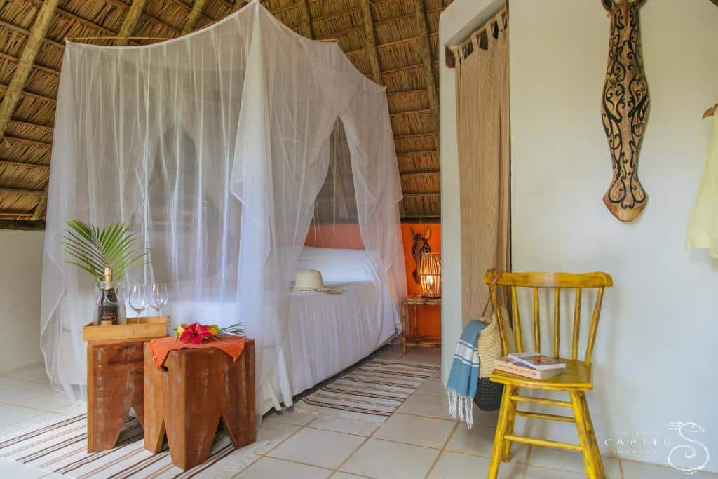 Uma cama com uma rede de dossel branca fica em um quarto de palha com móveis de madeira, tapetes e decoração simples, na Pousada Capitù em Imbassaí, para representar pousadas na Linha Verde na Bahia.