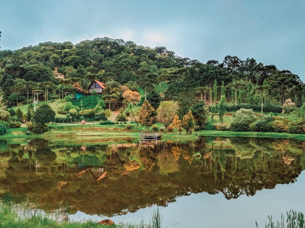 Casa em uma colina verde com árvores, refletida em um lago calmo sob um céu nublado.