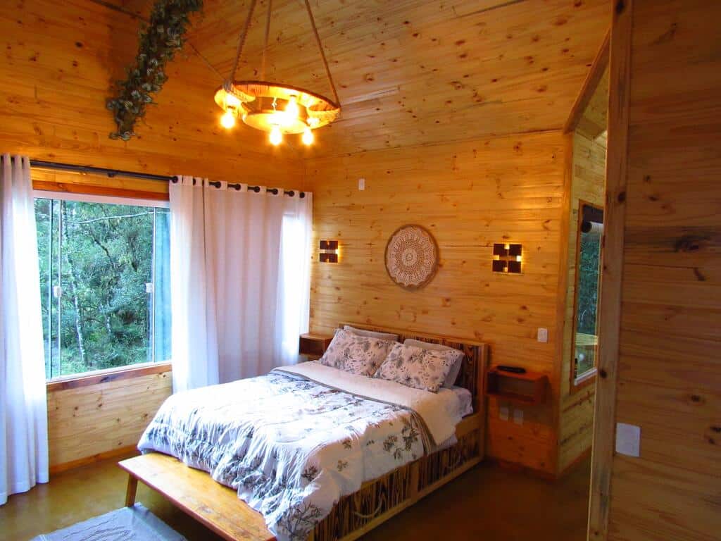 Quarto com paredes com painéis de madeira, uma cama com roupa de cama floral, uma janela com cortinas brancas e um lustre no alto.