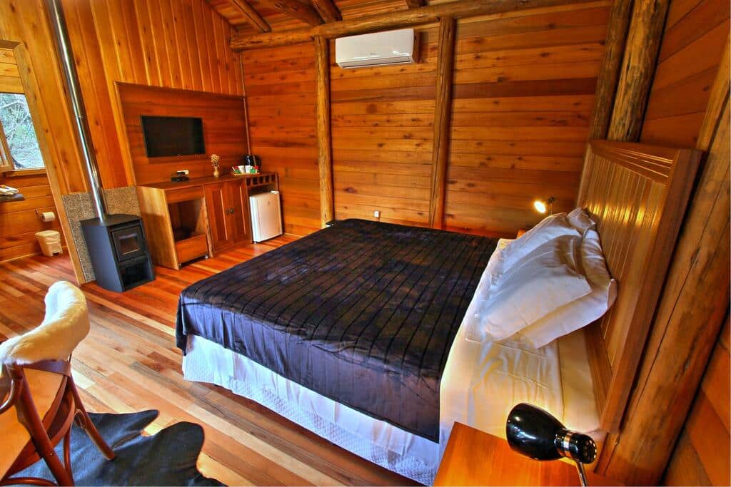 Um quarto em cabine de madeira com cama de casal, TV, escrivaninha, frigobar e paredes e pisos com painéis de madeira.