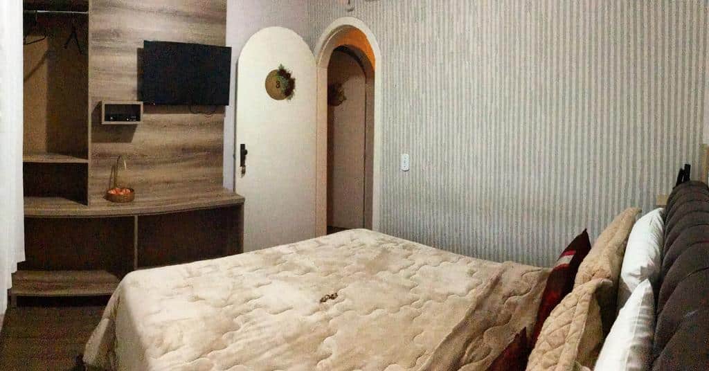 Uma cama bem arrumada fica de frente para uma TV na parede e um armário aberto em um quarto moderno com papel de parede listrado e porta em arco.