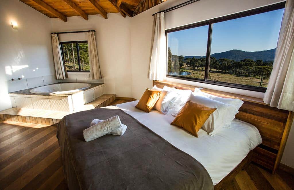 Um quarto com uma cama grande, travesseiros dourados, banheira de hidromassagem, piso de madeira e uma janela com vista para a montanha. Esta é uma das pousadas na serra catarinense.