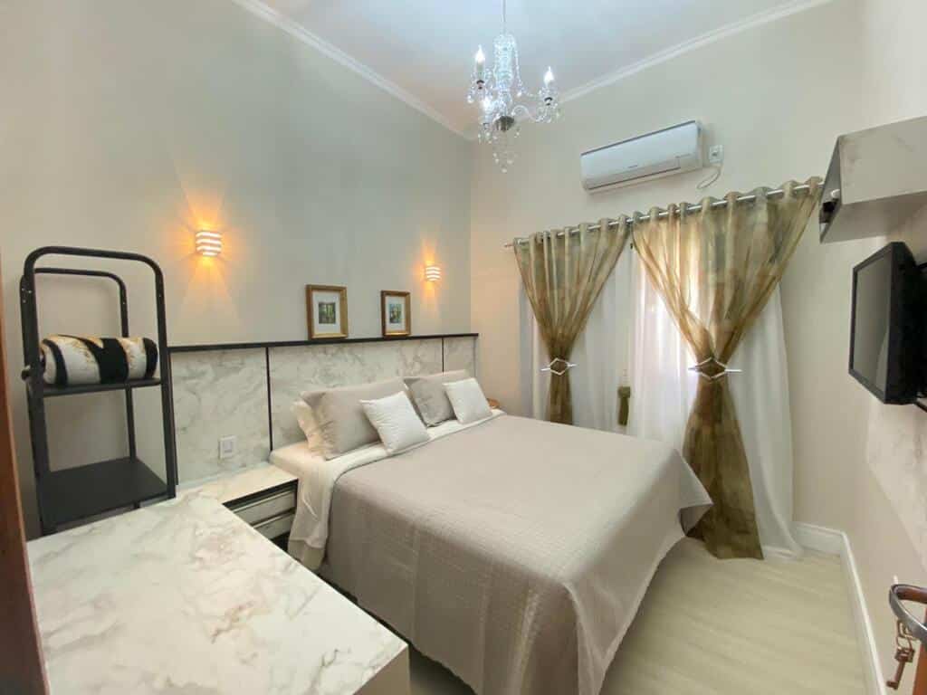 Uma cama bem arrumada em um pequeno quarto bege com luzes na parede, uma TV, cortinas e um lustre no alto.