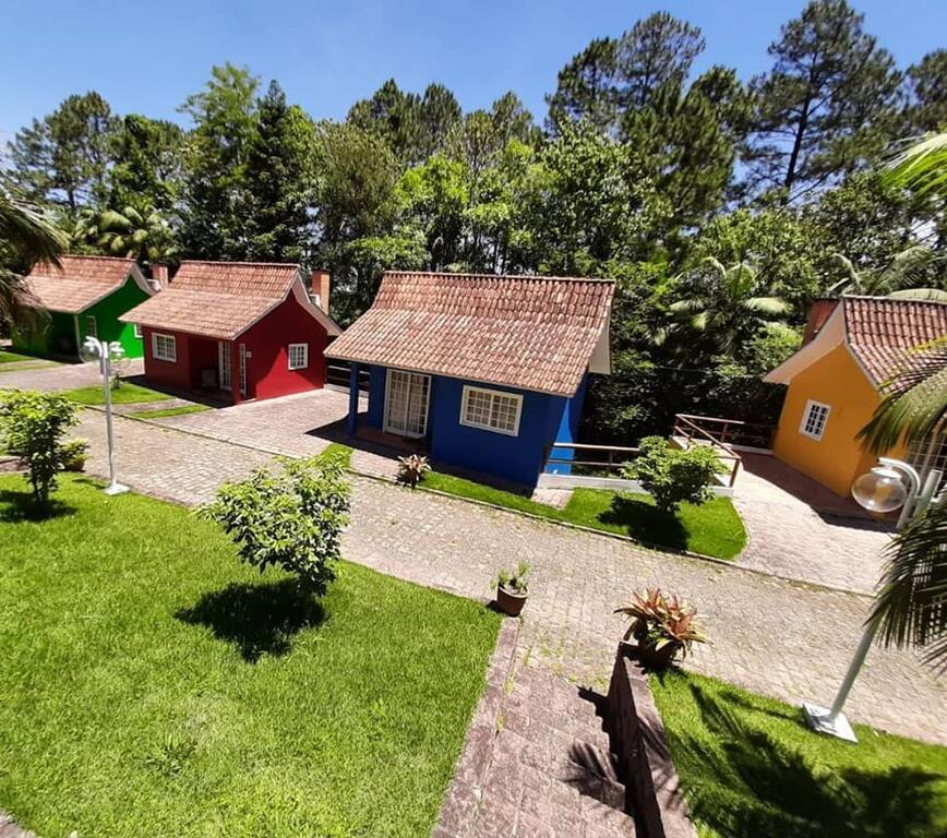 Três pequenas casas pintadas de vermelho, azul e amarelo ficam ao longo de um caminho de pedra cercado por árvores verdes e grama. Esta é uma das pousadas na serra catarinense.