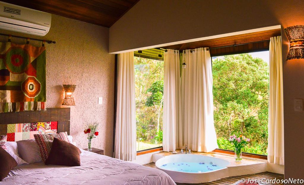 Um quarto com uma cama, decoração de parede e uma banheira redonda com grandes janelas com vista para as árvores verdes do lado de fora, na Pousada Serra do Luar em São Francisco Xavier, para representar pousadas no interior de SP para casal.