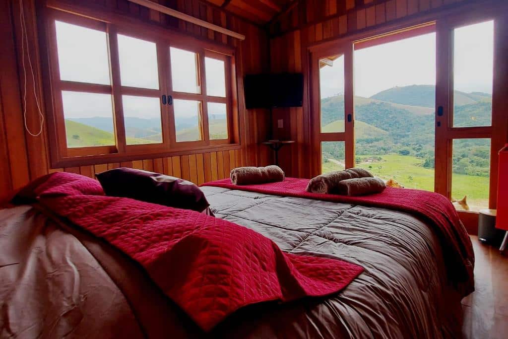 Quarto da Pousada Capim Limão em Monteiro Lobato com uma cama de casal de frente para um varanda com vista para as montanhas, além de algumas janelas, televisão e muitas almofadas em cima da cama