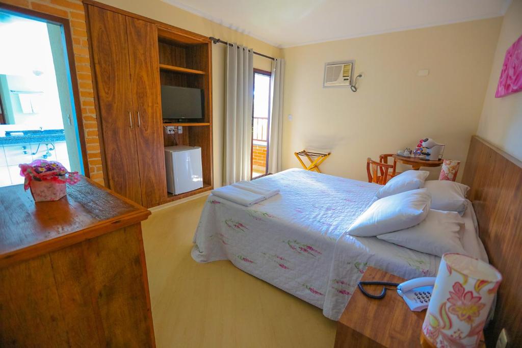 Um pequeno quarto com uma cama de casal, móveis de madeira, TV, frigobar, ar-condicionado e uma janela com cortinas, na Pousada Iguatiba em Paraibuna, para representar pousadas no interior de SP para casal.