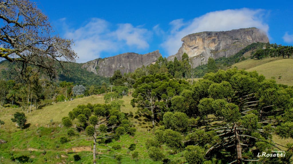 Colinas verdes com árvores espalhadas e montanhas rochosas ao fundo sob um céu azul com algumas nuvens, conhecido como Pedra do Baú em São Bento do Sapucaí.